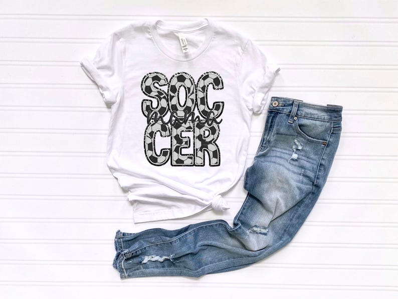 May include: White t-shirt with a black and white soccer ball design that says "Soccer Mama" on it. The shirt is paired with blue jeans.