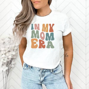 May include: White t-shirt with the text "In My Mom Era" in colorful retro-style lettering.