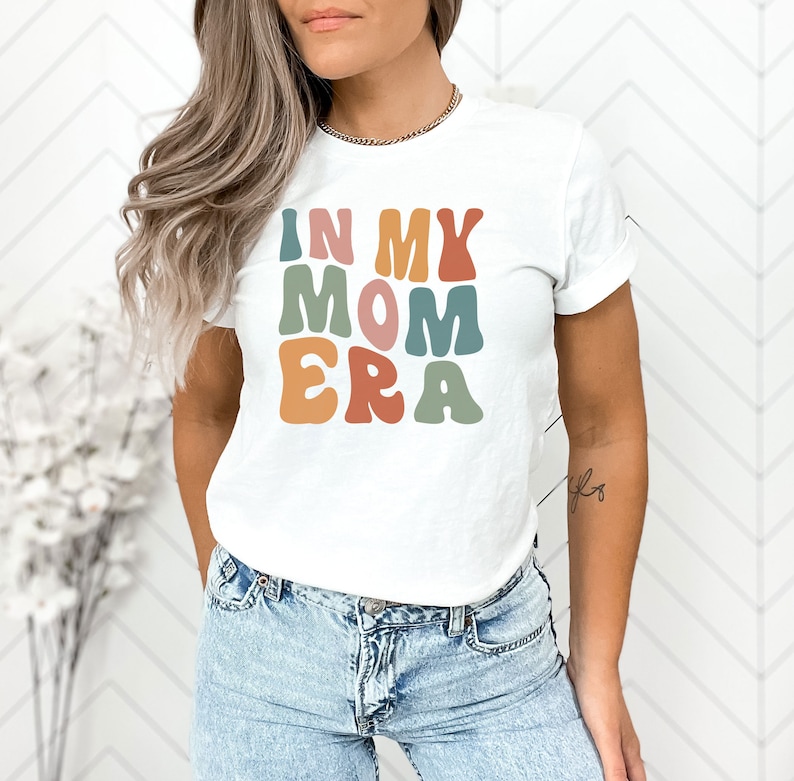 May include: White t-shirt with the text "In My Mom Era" in colorful retro-style lettering.