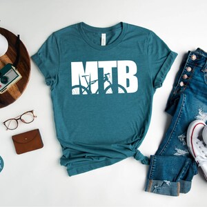 Puede incluir: Una camiseta de color azul teal jaspeado con un gr&aacute;fico blanco de una bicicleta de monta&ntilde;a con las letras MTB. La camiseta est&aacute; colocada sobre una superficie blanca con un par de pantalones vaqueros azules y zapatillas blancas.