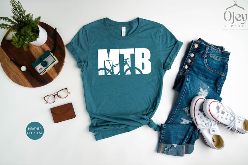 Puede incluir: Una camiseta de color azul teal jaspeado con un gr&aacute;fico blanco de una bicicleta de monta&ntilde;a con las letras MTB. La camiseta est&aacute; colocada sobre una superficie blanca con un par de pantalones vaqueros azules y zapatillas blancas.
