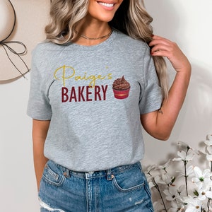 K&ouml;nnte beinhalten: Graues T-Shirt mit dem Text "Paige's Bakery" in goldenen Buchstaben und einer Grafik eines Schokoladen-Cupcakes.