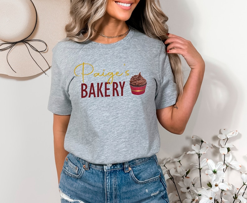K&ouml;nnte beinhalten: Graues T-Shirt mit dem Text "Paige's Bakery" in goldenen Buchstaben und einer Grafik eines Schokoladen-Cupcakes.