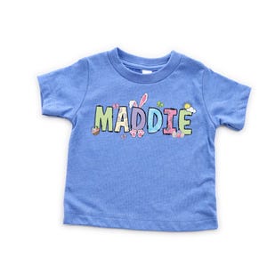 May include: A light blue toddler t-shirt with the name "MADDIE" in colorful, Easter-themed lettering. The design includes bunny ears, eggs, butterflies, and a sun, perfect for the spring season. The shirt is made of soft material.