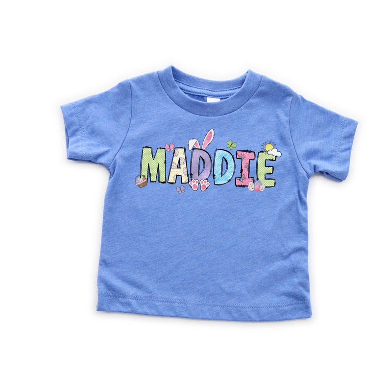 May include: A light blue toddler t-shirt with the name "MADDIE" in colorful, Easter-themed lettering. The design includes bunny ears, eggs, butterflies, and a sun, perfect for the spring season. The shirt is made of soft material.