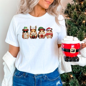 May include: White t-shirt with a graphic of four hamsters wearing Christmas hats and holding gifts. The hamsters are wearing a Santa hat, an elf hat, a reindeer hat, and a snowman hat.