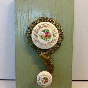 May include: A decorative wall hook with a floral design. The hook features a round, cream-colored porcelain element with a pink rose and green leaves, set in an ornate gold-colored metal frame. It is mounted on a light green wooden plaque.