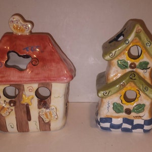 May include: Two ceramic birdhouse candle holders. The first is a single-story house with a red roof, brown and white stripes, and butterfly decorations. The second is a two-story house with a green roof, yellow and white checkered base, and sunflower decorations.