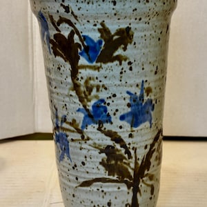 May include: A tall, speckled ceramic vase with a wide opening. The vase is off-white with brown and black speckles and features blue floral designs. The vase is approximately 8 inches tall.