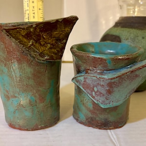May include: Two handmade ceramic vessels with a turquoise, brown, and gold color scheme. The taller vessel has a folded top, while the smaller one has a spout. The pottery has a rustic, textured appearance, suggesting a unique, artisanal craft.