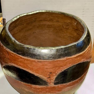 Puede incluir: Un cuenco redondo de cerámica hecho a mano con una amplia abertura. El cuenco presenta bandas de color naranja, negro y marrón, con un borde plateado. El diseño incluye formas oscuras estilizadas. El interior es de color marrón cálido.