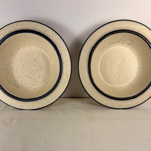 Könnte beinhalten: Zwei cremefarbene Keramikschalen mit gesprenkeltem Innenraum und dunkelblauen und schwarzen Rändern. Die Schalen sind rund und haben ein schlichtes, klassisches Design. Die Schalen dienen wahrscheinlich zum Servieren von Speisen.