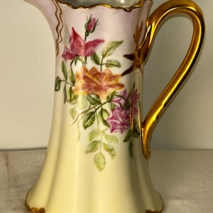 May include: A vintage porcelain pitcher with a lid, featuring a gold handle and trim. The pitcher is painted with colorful roses and green leaves. The body is a pale yellow, with a lavender rim and lid. The pitcher is approximately 8 inches tall.