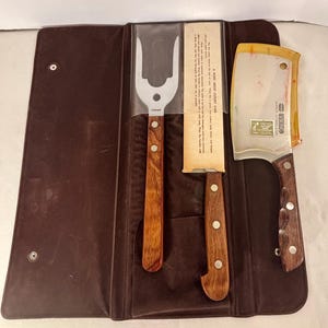 May include: A brown leatherette case contains a carving fork, a carving knife, and a cleaver. The utensils have wooden handles with silver rivets. The cleaver has a stainless steel blade with a yellow plastic guard. The case is open, displaying the contents.