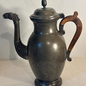 Puede incluir: Una tetera de peltre con acabado gris mate, que presenta un pico curvo moldeado con forma de cabeza de animal y detalles florales en la base. La tetera tiene una tapa abovedada con un pequeño remate y un asa de madera curvada.