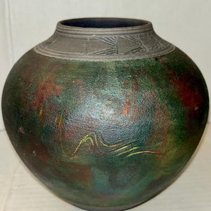 May include: A handcrafted, round ceramic vase with a textured, iridescent finish. The vase features a dark green base with red and gold accents. The rim is a darker gray with a geometric pattern. A unique decorative piece.