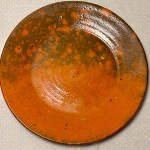 May include: A round ceramic plate featuring a glossy glaze with a gradient of vibrant orange at the bottom transitioning into a dark, mottled olive green at the top. The surface displays subtle circular texture marks and small dark speckles.