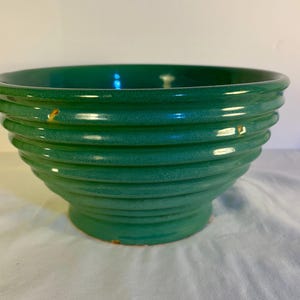 May include: A green ceramic bowl with a ribbed design. The bowl has a wide opening and a rounded base. The exterior is a glossy green color. The bowl is likely used for serving food or as a decorative piece.