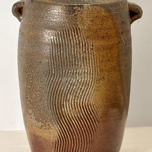 Pode incluir: Um vaso de cerâmica castanho artesanal com uma superfície texturizada e um design ondulado esculpido na frente. O vaso tem duas pequenas alças e mede aproximadamente 25 cm de altura.