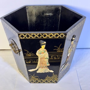 May include: Black and gold hexagonal decorative container with Asian-inspired artwork. The container features a black background with gold accents and images of figures in traditional attire. It has a brass handle on the side.