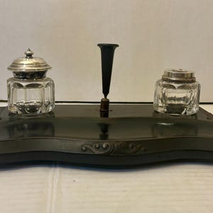 May include: Antique black inkwell set with two glass ink pots with silver lids and a black pen holder. The inkwell set is on a black base with decorative edges. A classic desk accessory.