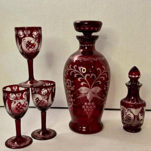 May include: A set of ruby red glassware, including three small wine glasses, a decanter with a stopper, and a small bottle. Each piece is etched with a white floral and grape design. The decanter is approximately 10 inches tall.