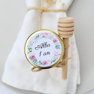 Peut inclure: Un petit pot avec un couvercle doré et une étiquette florale avec l'inscription "Alba 1 an". Un bâtonnet à miel en bois et une serviette blanche sont également visibles. Le pot est attaché avec de la ficelle.
