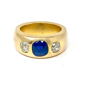 Può includere: Un anello d'oro con uno zaffiro blu e due diamanti. L'anello è incastonato in una fascia larga.