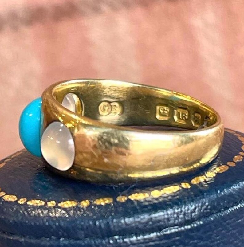 K&ouml;nnte beinhalten: Ein goldener Ring mit einem T&uuml;rkis- und einem wei&szlig;en Stein, der in einer Zargenfassung gefasst ist. Der Ring ist auf der Innenseite der Ringschiene mit den Buchstaben "GS" und "9ct" gekennzeichnet.