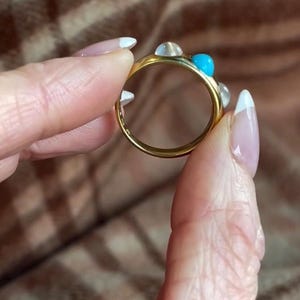 K&ouml;nnte beinhalten: Ein goldener Ring mit einem t&uuml;rkisfarbenen Stein und wei&szlig;en Steinen. Der Ring wird von einer Hand gehalten.