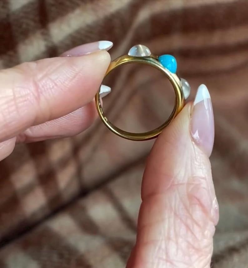 K&ouml;nnte beinhalten: Ein goldener Ring mit einem t&uuml;rkisfarbenen Stein und wei&szlig;en Steinen. Der Ring wird von einer Hand gehalten.