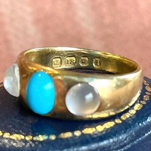 K&ouml;nnte beinhalten: Ein goldener Ring mit drei Steinen. Der mittlere Stein ist t&uuml;rkisfarben, die beiden &auml;u&szlig;eren Steine sind wei&szlig;. Der Ring ist mit einer Krone und der Zahl 18 gekennzeichnet.