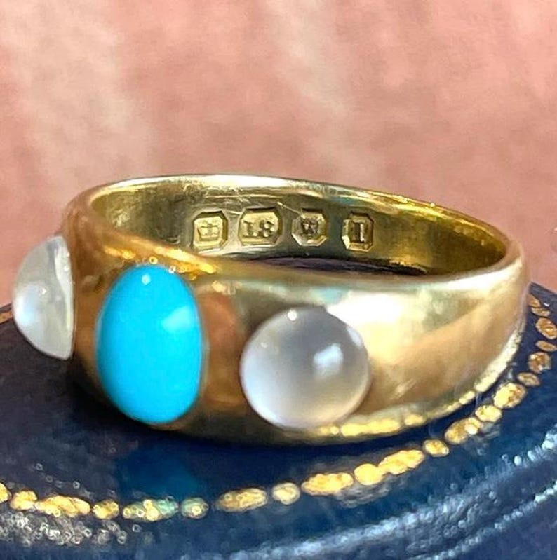 K&ouml;nnte beinhalten: Ein goldener Ring mit drei Steinen. Der mittlere Stein ist t&uuml;rkisfarben, die beiden &auml;u&szlig;eren Steine sind wei&szlig;. Der Ring ist mit einer Krone und der Zahl 18 gekennzeichnet.