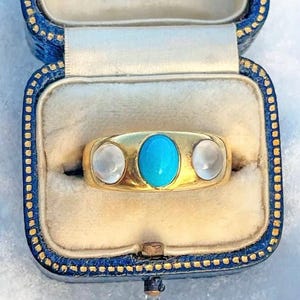 Può includere: Un anello d'oro con una pietra turchese e due pietre di luna. L'anello è in una scatola per anelli blu e oro.