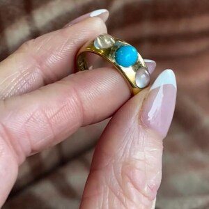 K&ouml;nnte beinhalten: Ein goldener Ring mit drei t&uuml;rkisfarbenen Steinen und vier wei&szlig;en Steinen. Der Ring befindet sich an einem Finger.