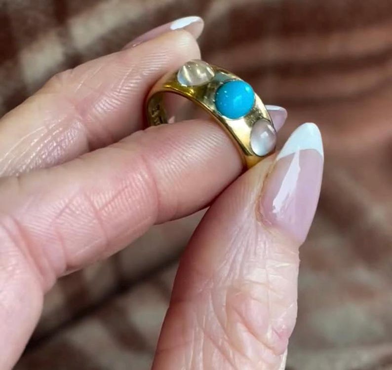 K&ouml;nnte beinhalten: Ein goldener Ring mit drei t&uuml;rkisfarbenen Steinen und vier wei&szlig;en Steinen. Der Ring befindet sich an einem Finger.