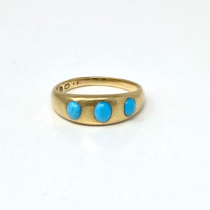 Può includere: Un anello d'oro con tre pietre ovali di turchese azzurro chiaro disposte sulla parte superiore. L'anello ha una fascia liscia e lucida e un design classico. L'anello è posto su uno sfondo bianco.