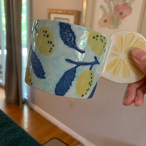 Pode incluir: Uma caneca de cer&acirc;mica com uma pega em forma de fatia de lim&atilde;o. A caneca &eacute; azul clara com lim&otilde;es amarelos e folhas azuis escuras. A pega tem a forma de uma fatia de lim&atilde;o com segmentos amarelos.