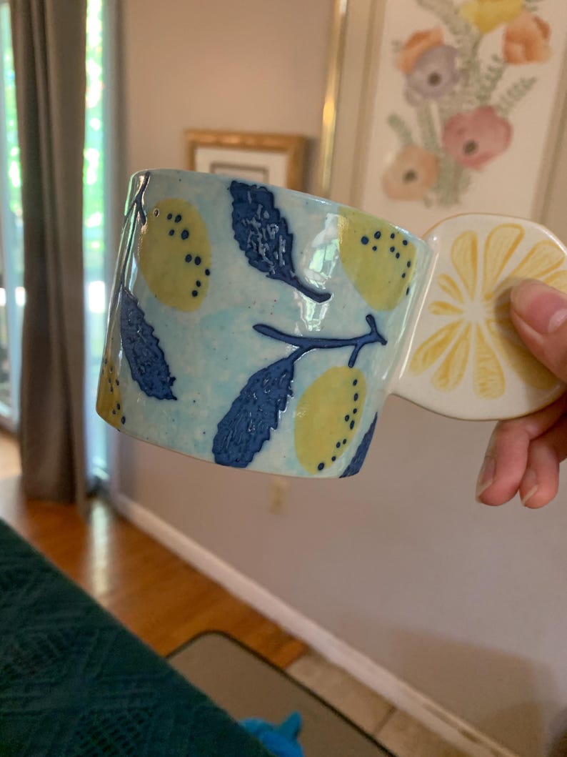 Pode incluir: Uma caneca de cer&acirc;mica com uma pega em forma de fatia de lim&atilde;o. A caneca &eacute; azul clara com lim&otilde;es amarelos e folhas azuis escuras. A pega tem a forma de uma fatia de lim&atilde;o com segmentos amarelos.