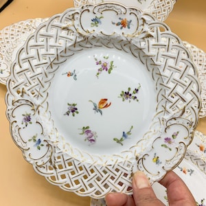 May include: White porcelain plates with intricate openwork lattice rims and delicate floral designs in various colors. The plates have gold trim and are partially visible, suggesting a set. The plates are likely for decorative or serving purposes.