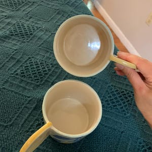 Pode incluir: Duas tigelas de sopa de cer&acirc;mica com al&ccedil;as. As tigelas s&atilde;o branco-sujas com uma al&ccedil;a amarelo p&aacute;lido e um desenho de listras azuis e amarelas ao redor da borda. As tigelas est&atilde;o sobre um cobertor verde-azulado texturizado.