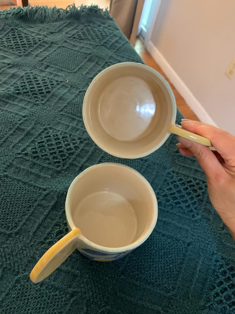 Pode incluir: Duas tigelas de sopa de cer&acirc;mica com al&ccedil;as. As tigelas s&atilde;o branco-sujas com uma al&ccedil;a amarelo p&aacute;lido e um desenho de listras azuis e amarelas ao redor da borda. As tigelas est&atilde;o sobre um cobertor verde-azulado texturizado.