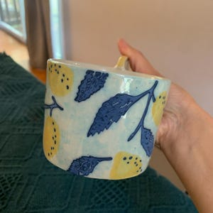 Pode incluir: Uma caneca de cer&acirc;mica com fundo azul claro, adornada com lim&otilde;es amarelos e folhas azuis. A caneca tem uma pequena al&ccedil;a e uma forma ligeiramente quadrada. O design &eacute; pintado &agrave; m&atilde;o, dando-lhe um visual art&iacute;stico &uacute;nico.