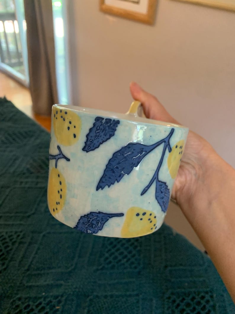 Pode incluir: Uma caneca de cer&acirc;mica com fundo azul claro, adornada com lim&otilde;es amarelos e folhas azuis. A caneca tem uma pequena al&ccedil;a e uma forma ligeiramente quadrada. O design &eacute; pintado &agrave; m&atilde;o, dando-lhe um visual art&iacute;stico &uacute;nico.