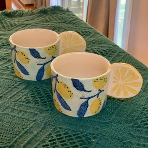 Pode incluir: Duas canecas de cer&acirc;mica com uma al&ccedil;a em forma de fatia de lim&atilde;o. As canecas s&atilde;o brancas com um fundo azul claro e apresentam lim&otilde;es amarelos e folhas azuis. As canecas est&atilde;o sobre uma superf&iacute;cie texturizada verde-azulado.