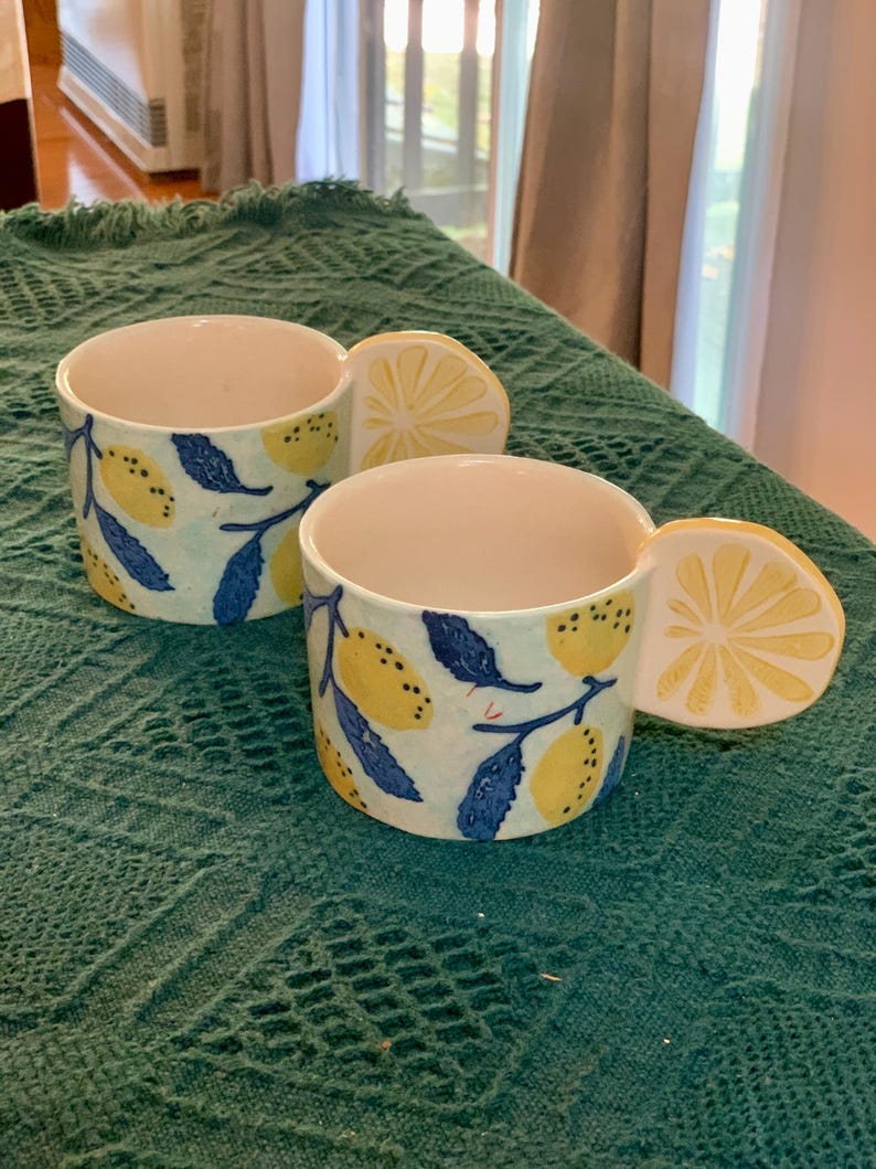 Pode incluir: Duas canecas de cer&acirc;mica com uma al&ccedil;a em forma de fatia de lim&atilde;o. As canecas s&atilde;o brancas com um fundo azul claro e apresentam lim&otilde;es amarelos e folhas azuis. As canecas est&atilde;o sobre uma superf&iacute;cie texturizada verde-azulado.
