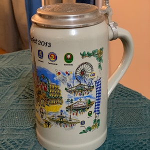 May include: A ceramic beer stein with a metal lid and handle. The stein is cream-colored and features colorful illustrations of a festival, including a Ferris wheel and beer barrels. The text "Oktoberfest 2013" is printed on the front.
