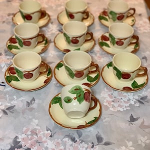 Könnte beinhalten: Ein Set aus 10 Vintage-Teetassen und Untertassen mit einem Apfeldesign in Braun, Grün und Rot. Die Tassen und Untertassen sind weiß mit einer cremefarbenen Glasur.