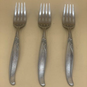 Pode incluir: Três garfos de prata com desenhos ornamentados nas alças. Os garfos estão dispostos em fila sobre uma superfície bege.