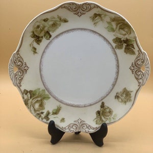Peut inclure: Une assiette de service en porcelaine blanche avec un bord festonné et un motif floral composé de roses vertes et de feuilles brunes. L'assiette a un bord surélevé et une bordure décorative.